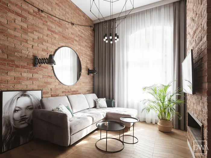 Loftowy salon z cegłą, okrągłym lustrem i szarą sofą pikowaną - apartament w Krakowie 22m2